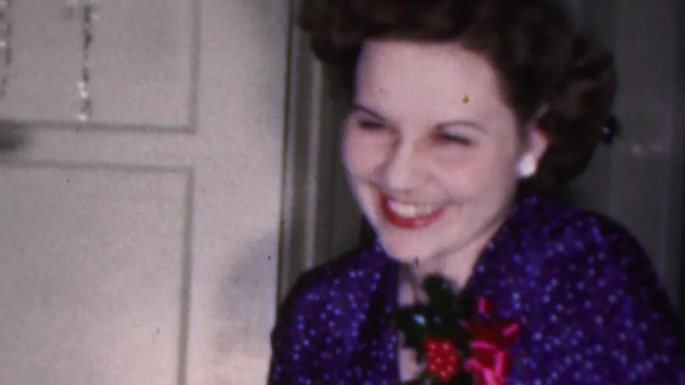 A close 8mm frame shows the woman laughing or smiling broadly in festive purple clothing with a red-and-green brooch, framed beside a white paneled door.