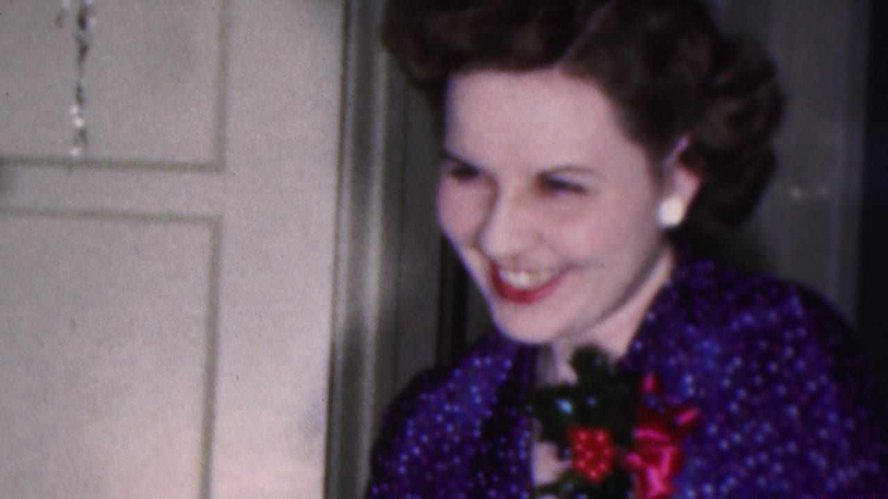 In a 1953 Cleveland home-movie frame, a smiling woman with styled dark hair wears a blue patterned dress, white earrings, red lipstick, and a corsage near a white paneled door.