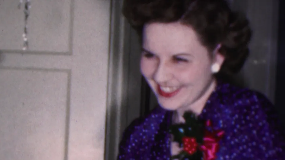 In a 1953 Cleveland home-movie frame, a smiling woman with styled dark hair wears a blue patterned dress, white earrings, red lipstick, and a corsage near a white paneled door.