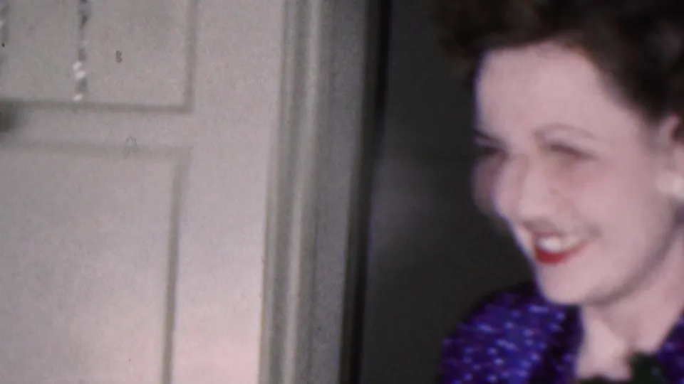 A soft-focus 1953 Cleveland frame shows the woman in partial profile, smiling beside a white paneled door with her corsage and blue patterned dress partly visible.