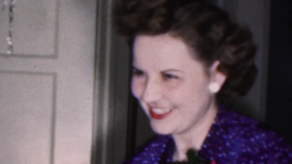 The woman smiles while looking off-camera, showing red lipstick, white earrings, styled dark hair, and a blue patterned dress beside a white interior door in the 1953 Cleveland footage.