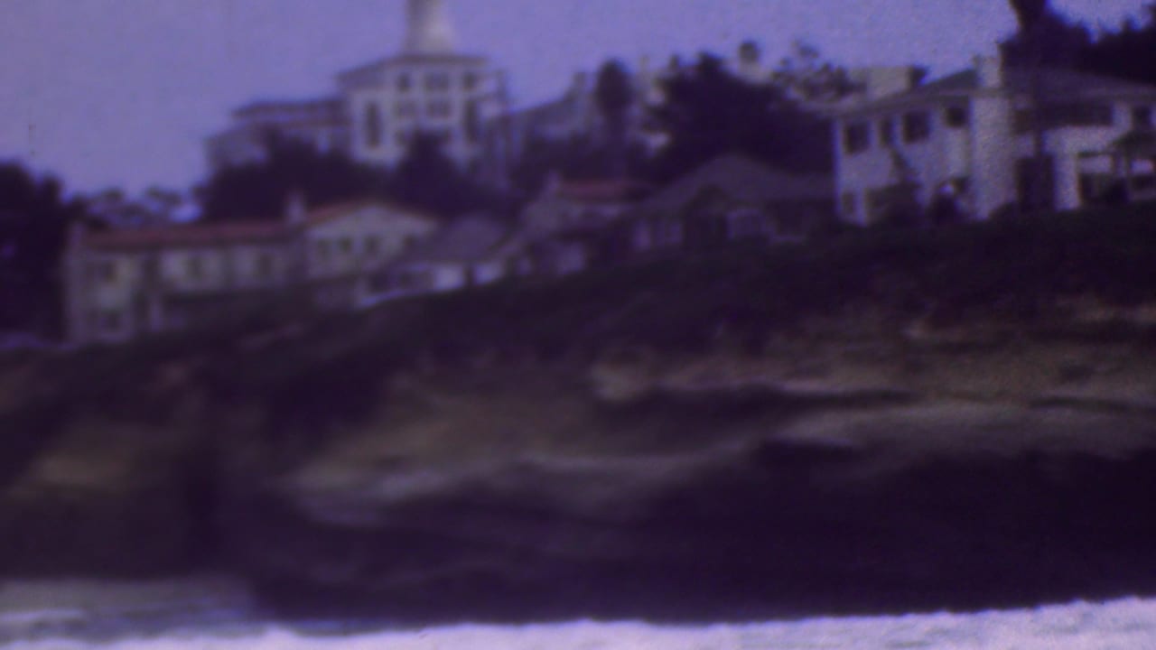 Soft 8mm view of houses and larger structures along a bluff, with dark shoreline rock and surf in the lower frame.