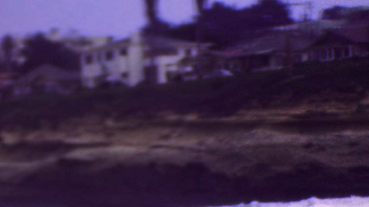Home-movie frame showing light-colored coastal houses, trees, and layered rock ledges near the waterline in the Cleveland-identified clip.