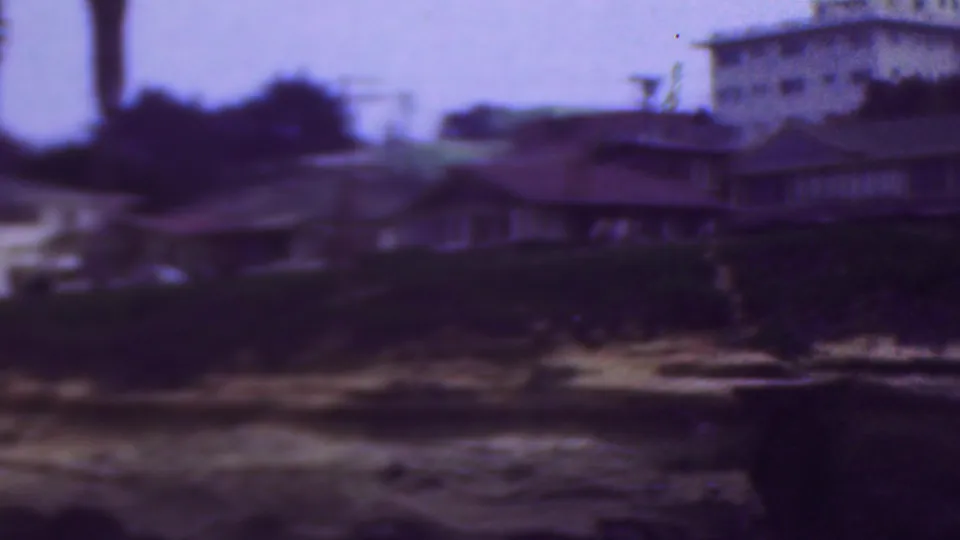 1953 8mm frame from the supplied Cleveland, Ohio metadata showing blurred beach houses and a larger building above a sandy shoreline and dark water.
