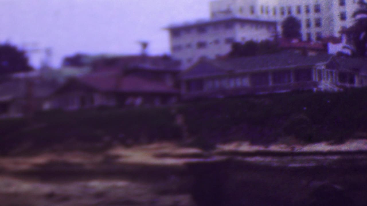 Soft 8mm view of a coastal residential slope with low houses, vegetation, a pale multi-story building, sand, and water in the foreground.
