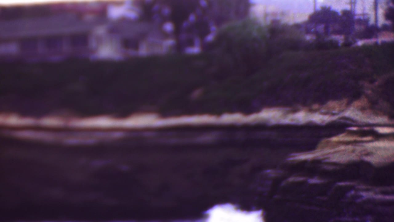1953 Cleveland, Ohio 8mm frame showing waves along a dark rocky shoreline beneath soft-focus houses, trees, and pale bluff edges.