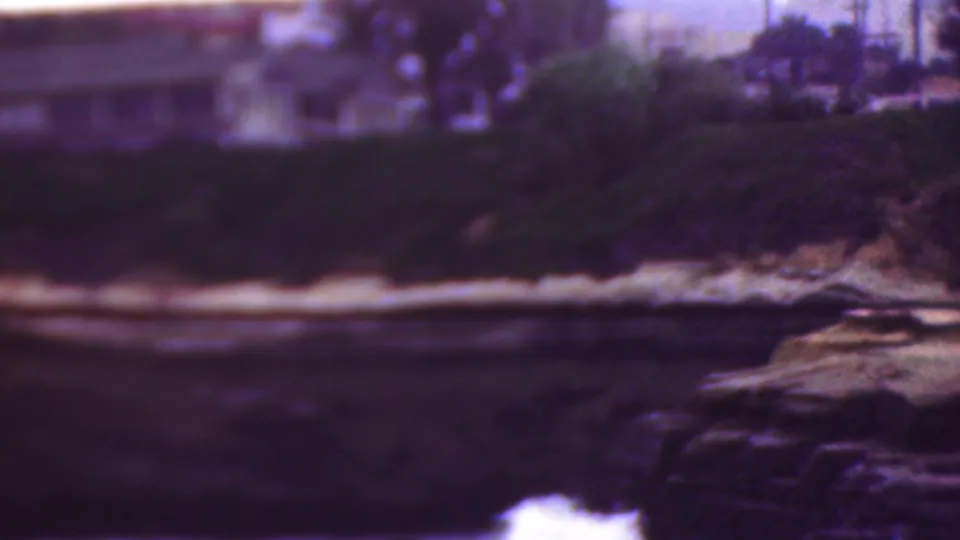 1953 Cleveland, Ohio 8mm frame showing waves along a dark rocky shoreline beneath soft-focus houses, trees, and pale bluff edges.