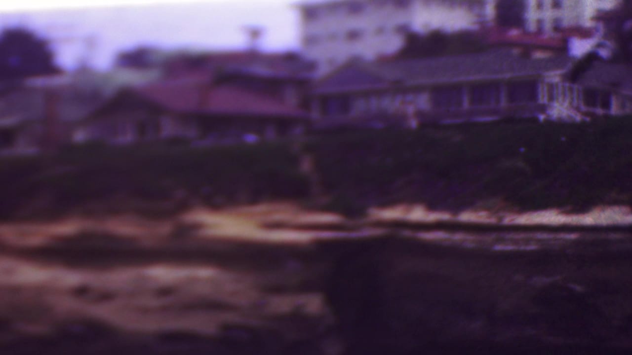 Blurred houses and trees above a dark rocky beach with foamy water.