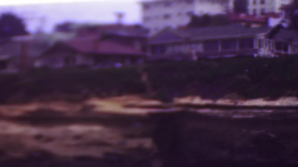1953 Cleveland, Ohio home-movie frame with residential buildings and trees above a dark, rocky shoreline where small waves break into foam.