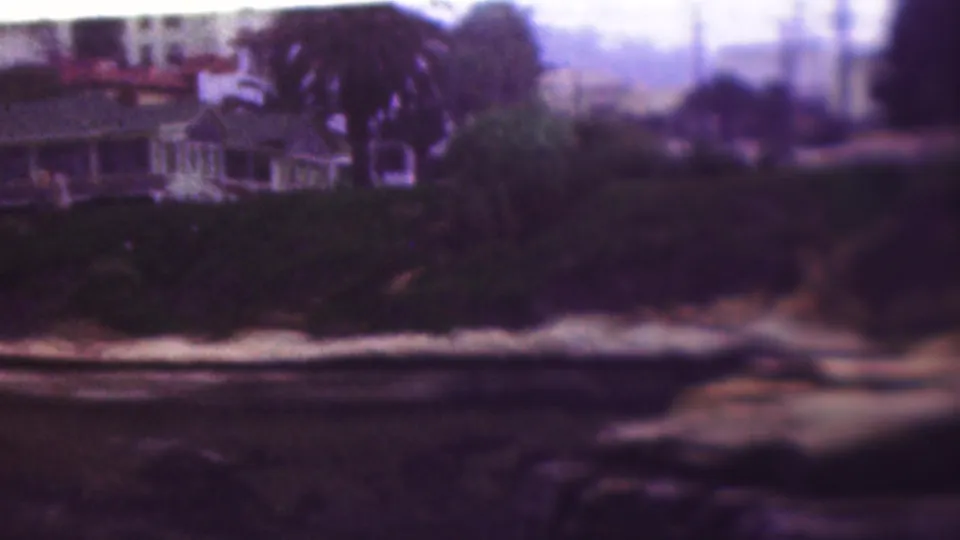1953 Cleveland, Ohio frame showing a cliffside residence, palm-like trees or tall plantings, dense shrubs, and pale surf along the rocky waterline.
