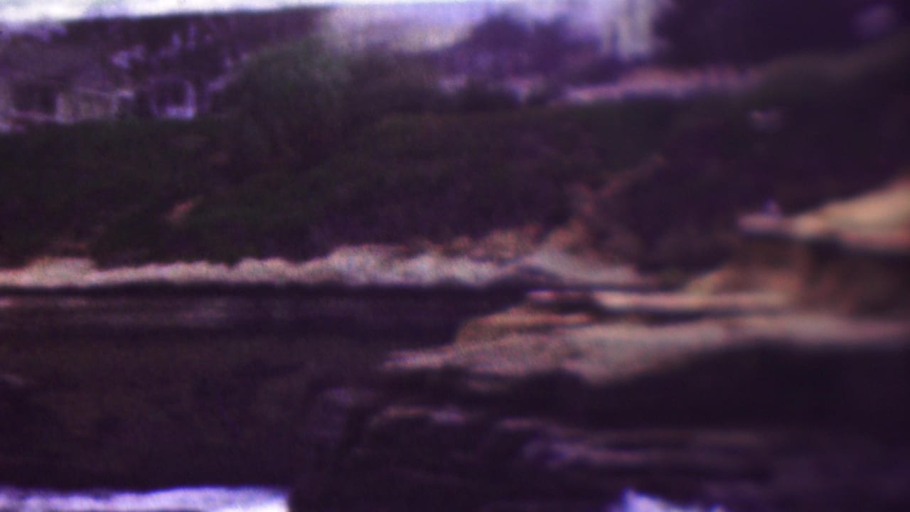 1953 8mm home-movie frame from the supplied Cleveland, Ohio clip showing a rocky shoreline or cliff edge above dark water, with vegetation and pale railings in the distance.