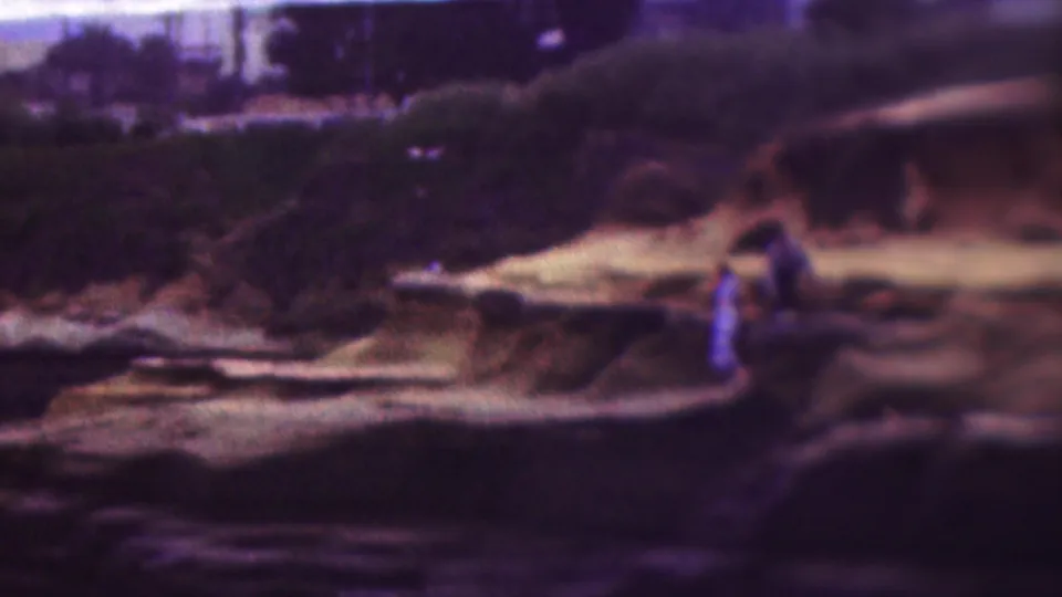 1953 8mm home-movie frame from the supplied Cleveland, Ohio clip showing two distant adults near a pale rock shelf beside a dark, rugged shoreline.
