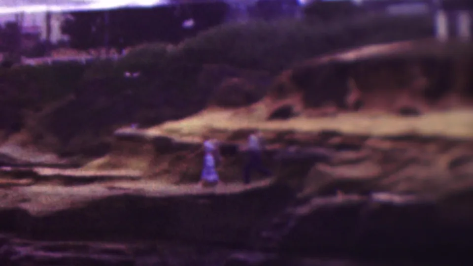 1953 8mm home-movie frame from the supplied Cleveland, Ohio clip showing two adults walking along a rocky ledge near water, with shrubs and pale stone behind them.