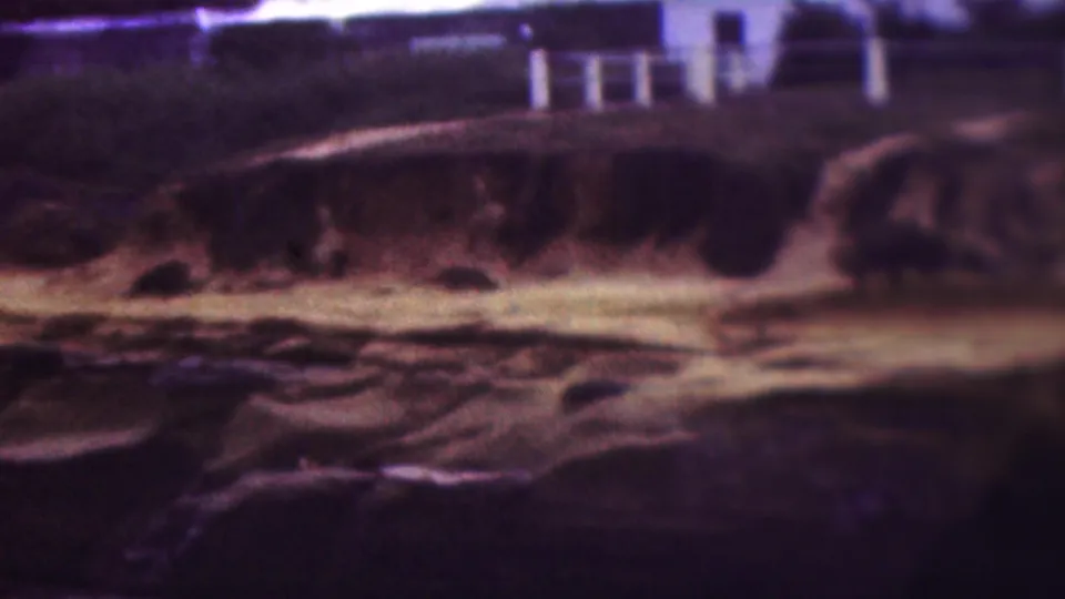 1953 8mm home-movie frame from the supplied Cleveland, Ohio clip showing pale rock ledges, dark water, vegetation, and white railings along the upper shoreline area.