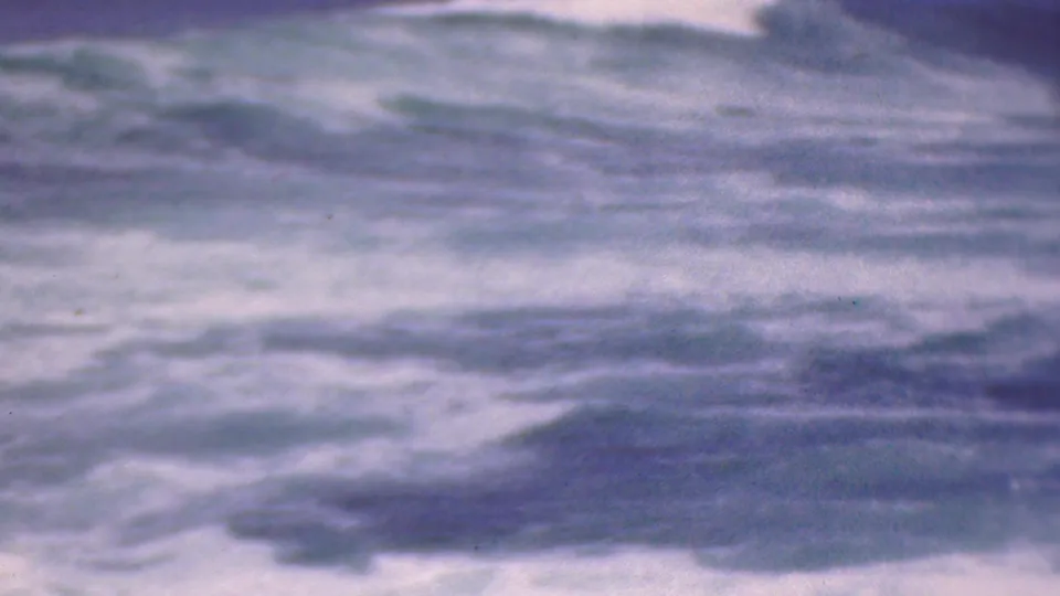 Close, soft-focus view of rough foamy water with darker wave bands and aged blue-purple 8mm film texture.