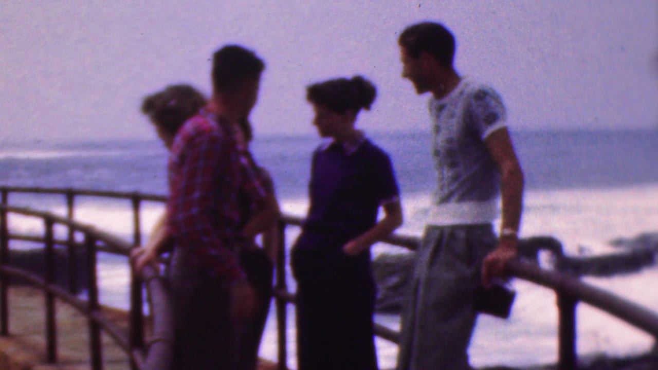 Four teenagers in 1953 home-movie footage stand near a boardwalk railing with ocean surf behind them, including a red plaid shirt, dark dress, and light patterned T-shirt.