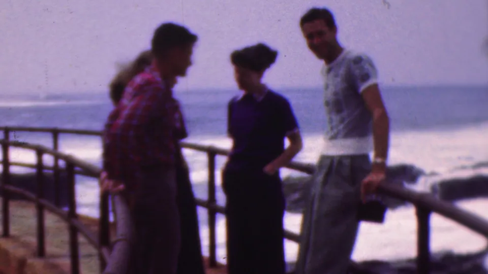 A 1953 8mm frame shows teenagers talking beside a seaside railing, with a young man in a red plaid shirt, a young woman in a dark dress, and a young man in a light patterned T-shirt.