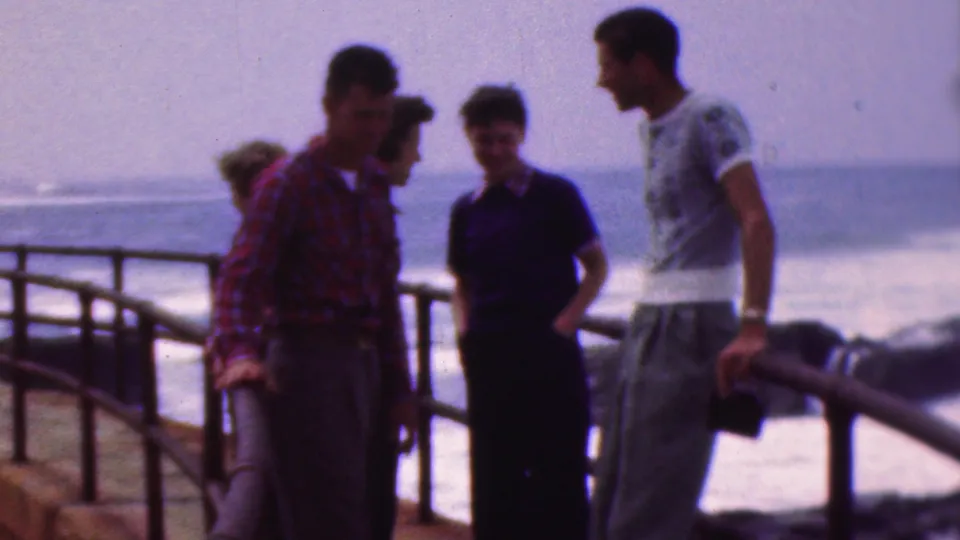 Four teenagers appear in a grainy 1953 home-movie frame by a seaside railing, with ocean waves, casual clothing, and a hazy horizon visible behind them.