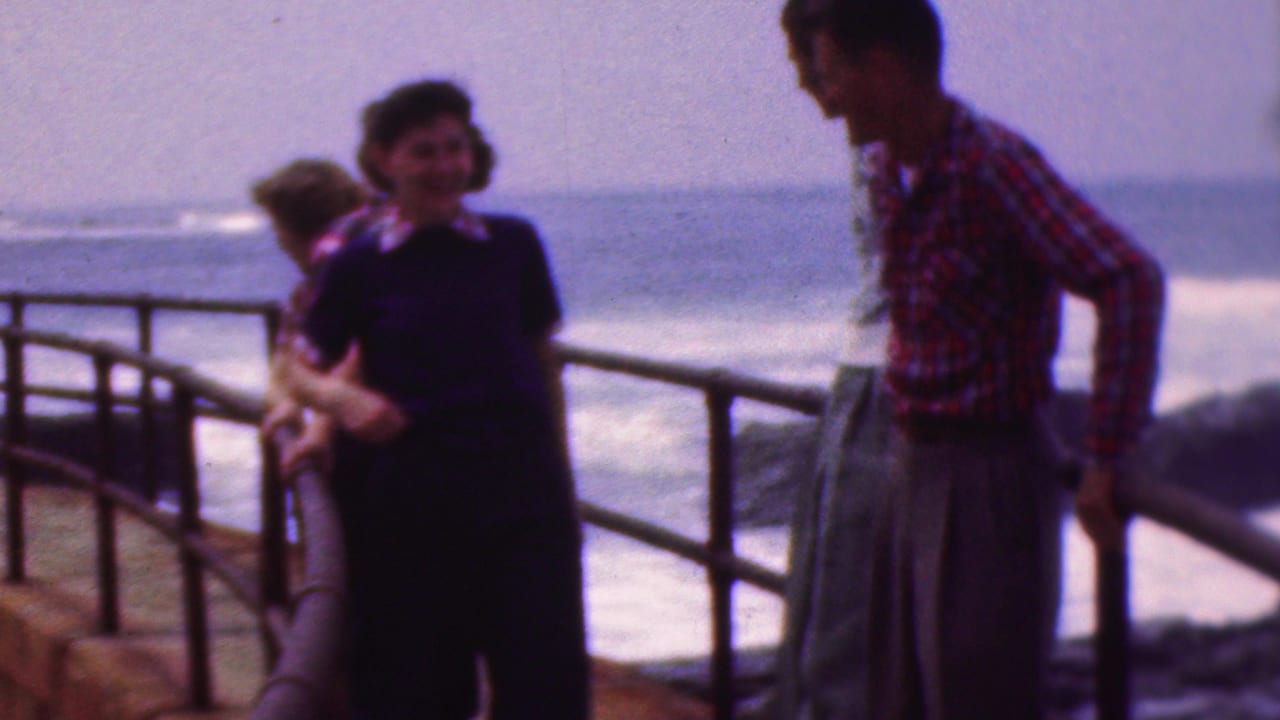 A woman in a dark dress with light trim and a man in a red plaid shirt face each other near a waterfront railing in the 1953 Cleveland home-movie clip.