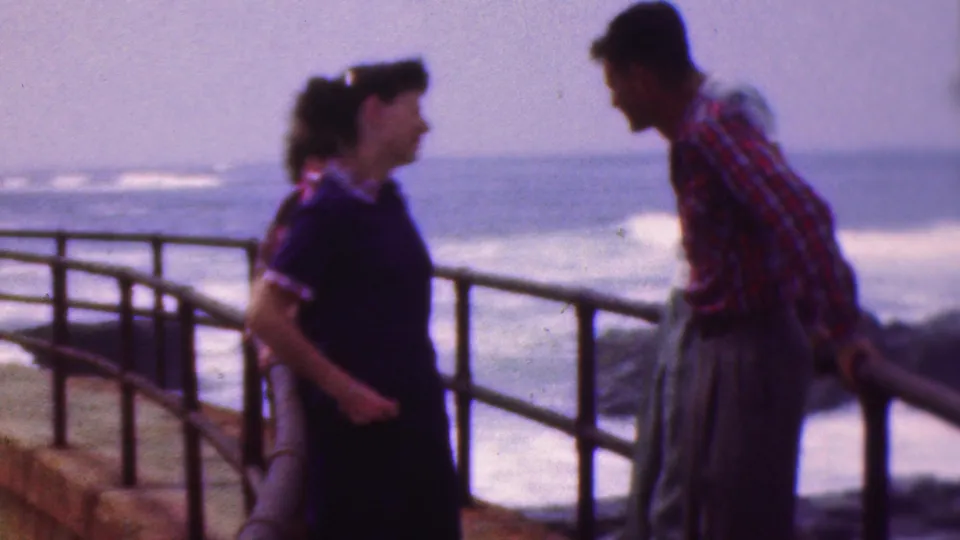 The handheld 8mm view shows a woman and a man talking by the curved pier railing, with blurred waves and shoreline behind them.