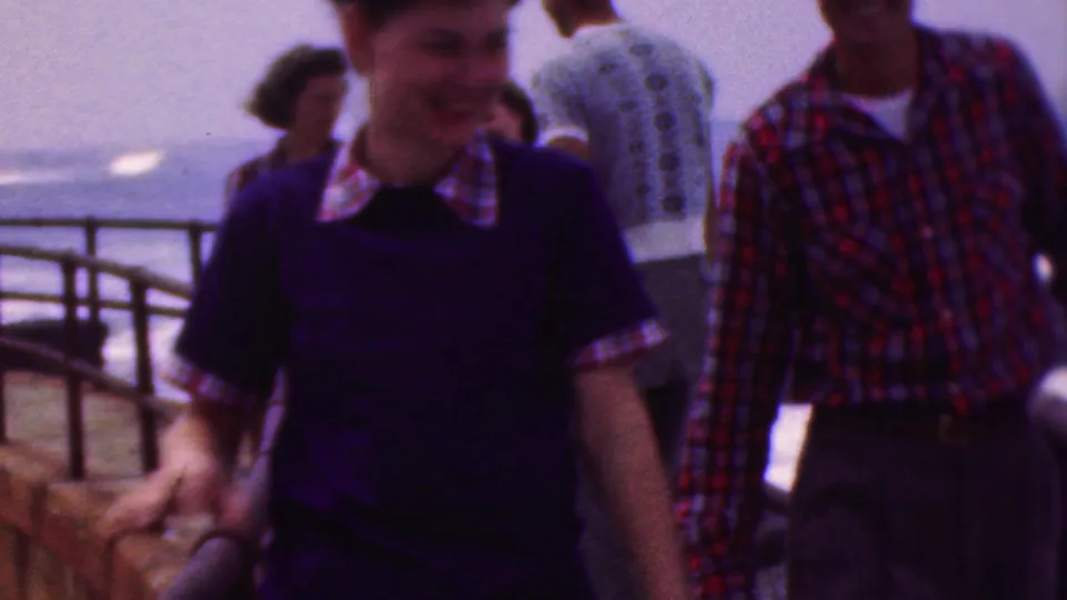 A young woman in a dark dress passes close to the camera as other adults stand near the curved railing and open water behind her.
