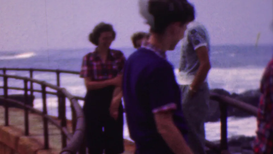 A woman in a dark outfit crosses the foreground as the remaining friends stand along the waterside railing, with open water and waves in the background.