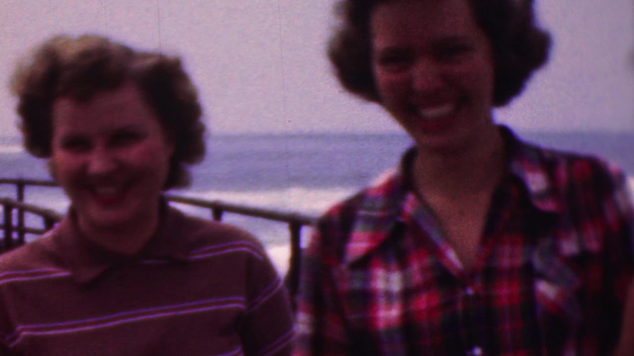 Three young women in striped and plaid tops smile and laugh near metal railings, with open water and a hazy horizon behind them in the supplied Cleveland, Ohio home-movie setting.