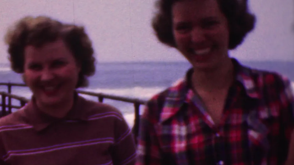 Three young women in striped and plaid tops smile and laugh near metal railings, with open water and a hazy horizon behind them in the supplied Cleveland, Ohio home-movie setting.