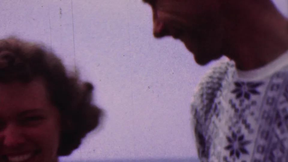 A laughing young woman appears at the edge of the frame while a young man in a patterned sweater looks downward beside her against a bright waterfront background.