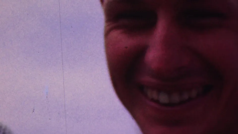 An extreme close-up catches a young woman smiling into the handheld camera, with little background detail beyond pale sky and soft film grain.