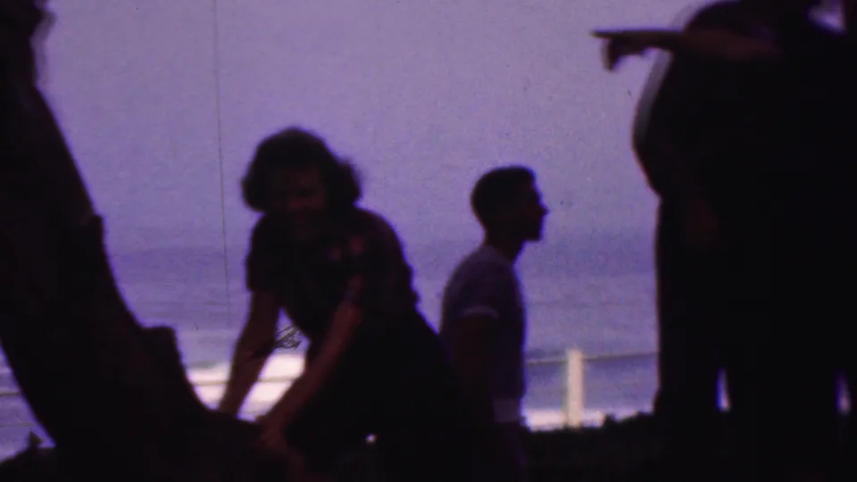 Several silhouetted adults, including a woman near a tree and a man in profile, stand by a railing with water beyond in a 1953 8mm home-movie frame attributed to Cleveland, Ohio.