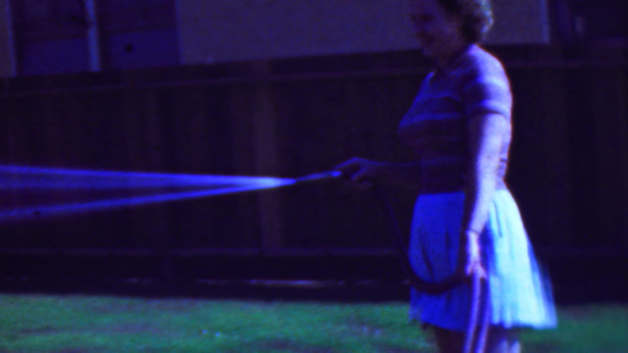 In a 1953 Cleveland home-movie frame, a woman in a striped top and light skirt stands on a lawn by a wooden fence while spraying water from a garden hose.