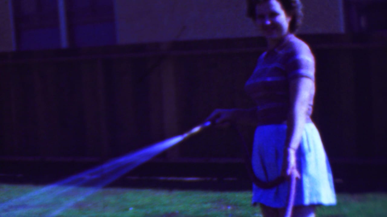 Woman looking toward the camera while directing a hose spray across the lawn.
