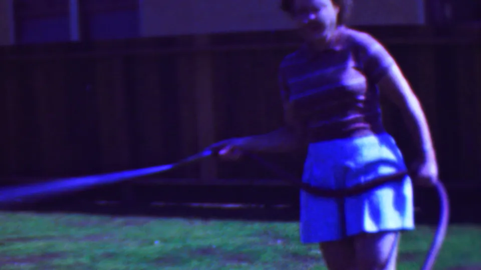 A closer 1953 home-movie view shows the woman with the hose looped at her side while the water stream extends across the lawn in front of a wooden fence.
