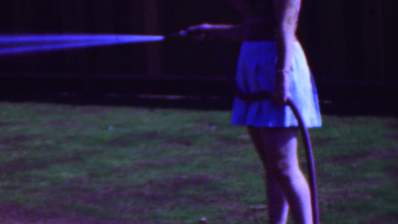 In 1953 Cleveland home-movie footage, a woman in a light blue skirt holds a hose while water sprays across a shaded grassy backyard.