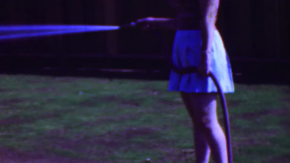 In 1953 Cleveland home-movie footage, a woman in a light blue skirt holds a hose while water sprays across a shaded grassy backyard.