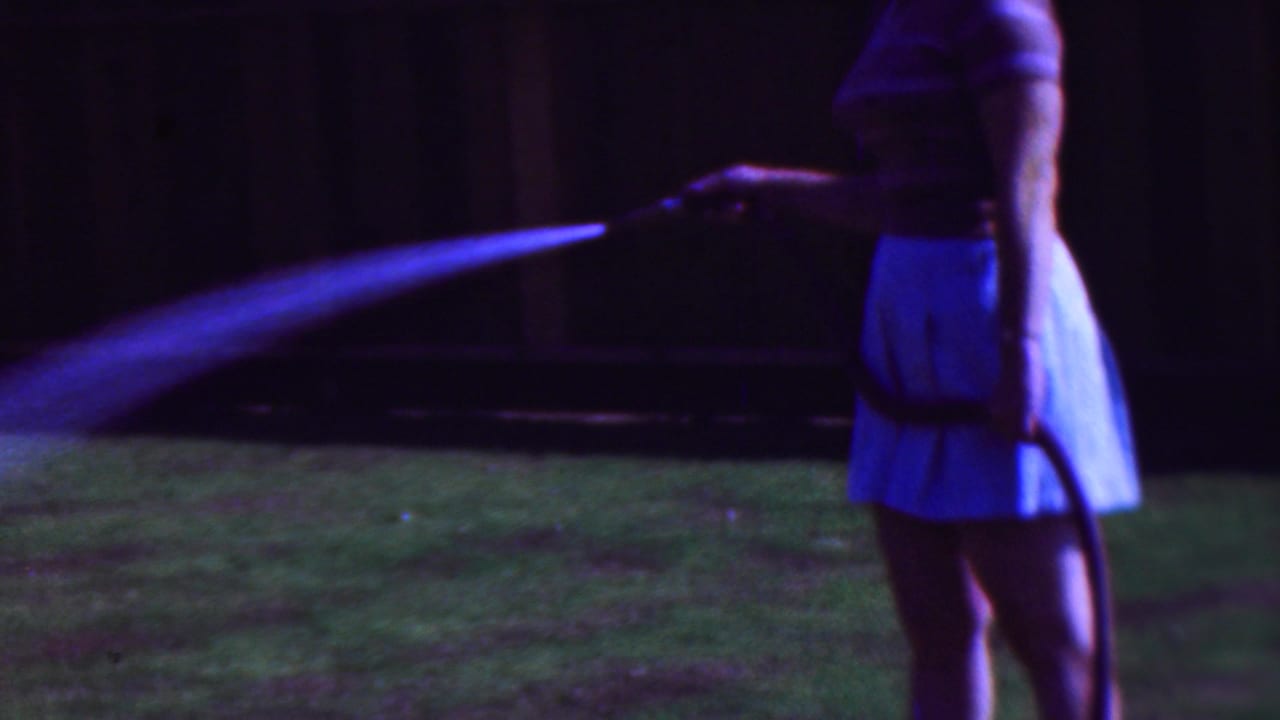Woman stands at the right edge holding a hose as water arcs across the lawn.