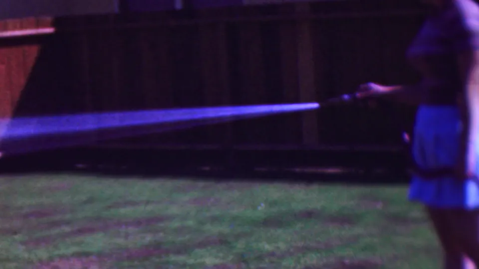 A soft-focus 8mm frame shows the woman in profile watering the lawn, with a pale post and wooden fence visible behind her.