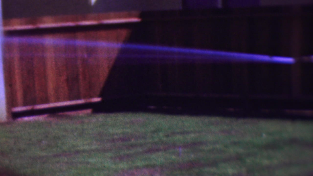 1953 Cleveland 8mm frame showing a purple-blue water stream arcing across a grassy backyard beside a wooden fence, with the woman mostly outside the right edge.