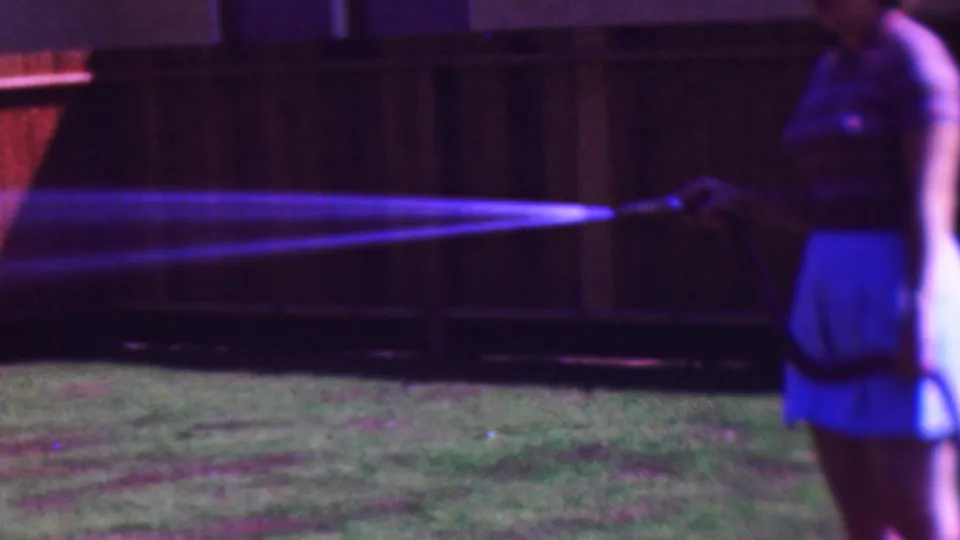 1953 Cleveland home-movie frame showing a woman in a light skirt standing at the right edge and directing a garden hose across a grassy backyard under artificial light.