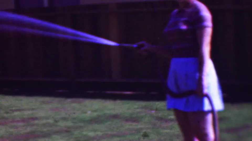 1953 Cleveland 8mm frame showing a woman on a backyard lawn holding a garden hose as water fans across the frame in a purple-blue cast.
