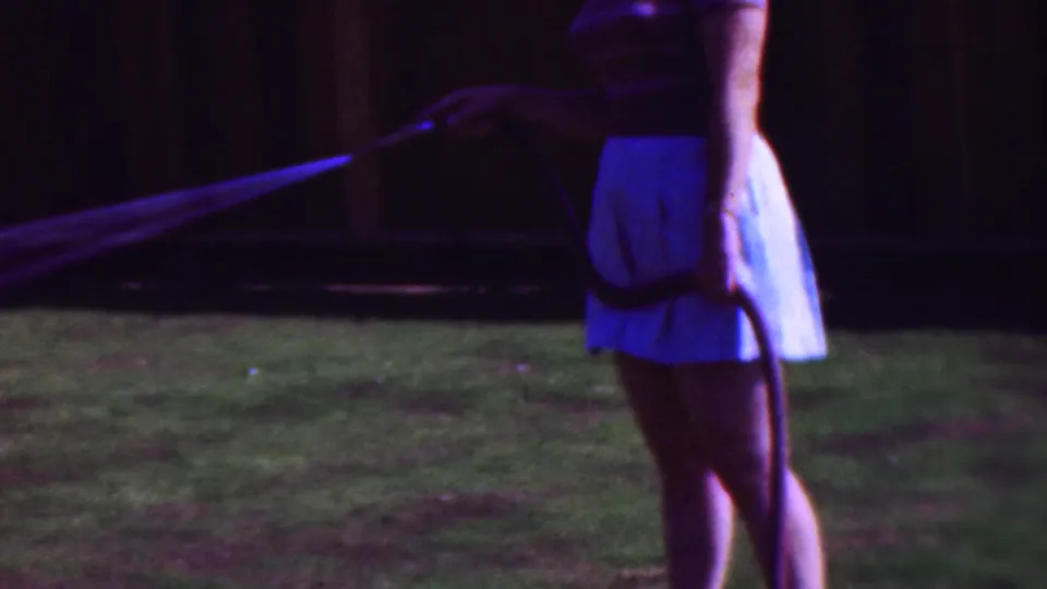 1953 Cleveland frame showing a woman in a light skirt and short-sleeved top holding a garden hose as water streams across a dark grassy backyard.