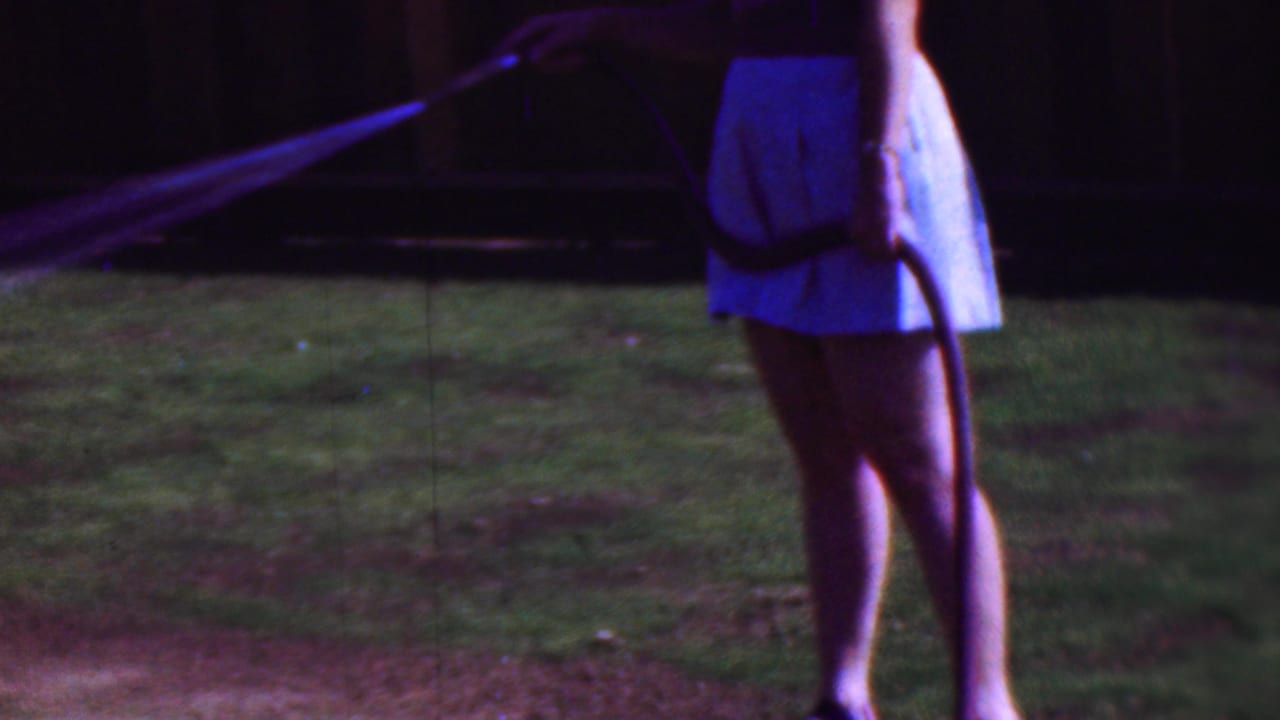 In this 1953 Cleveland home-movie frame, a woman in a light skirt and sandals stands on grass while directing a garden hose spray across the yard.