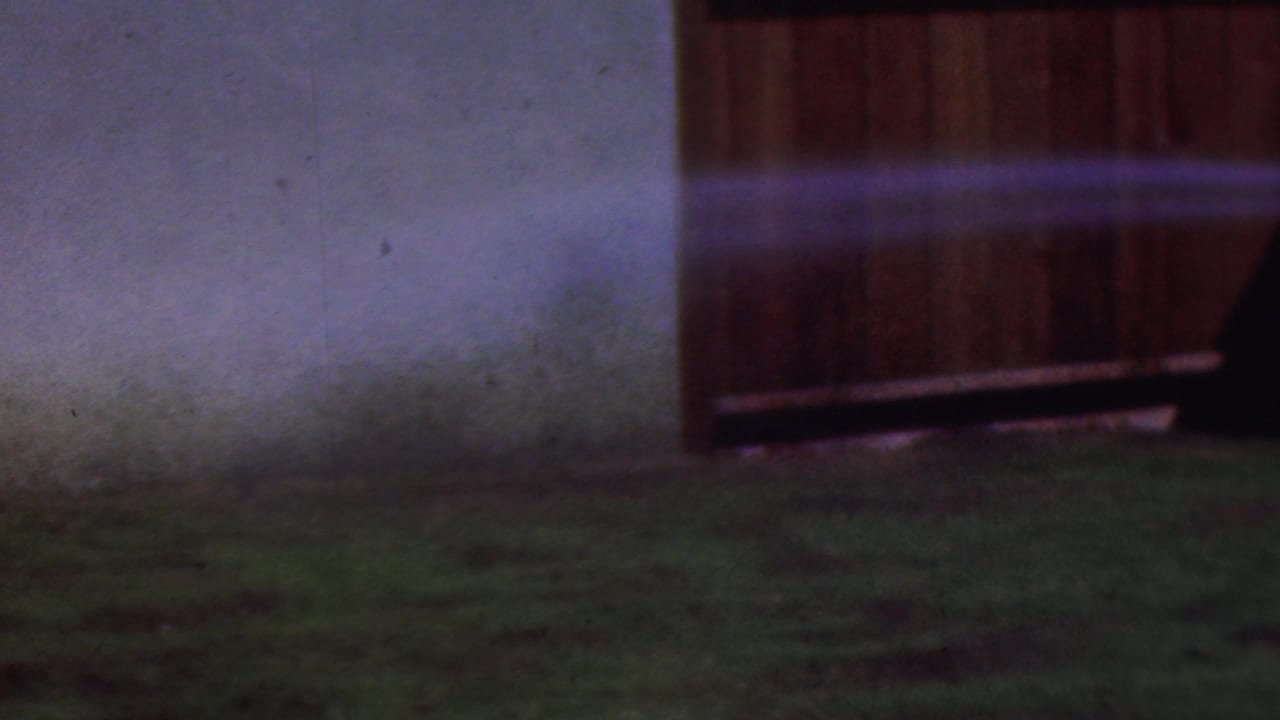 In a Cleveland backyard setting supported by the clip metadata, a hose stream crosses patchy grass toward a pale wall and a wooden fence, with soft 8mm color and motion blur.