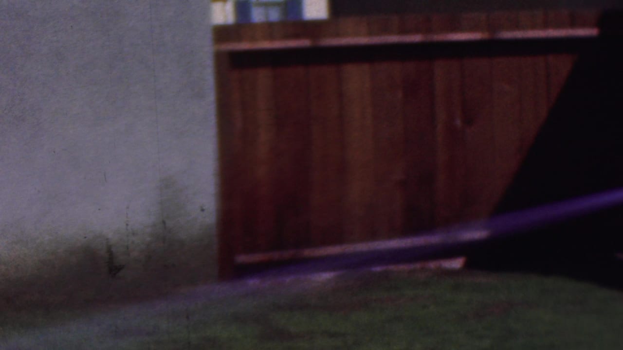 A hose stream sprays toward a wooden fence beside a pale wall and grass.