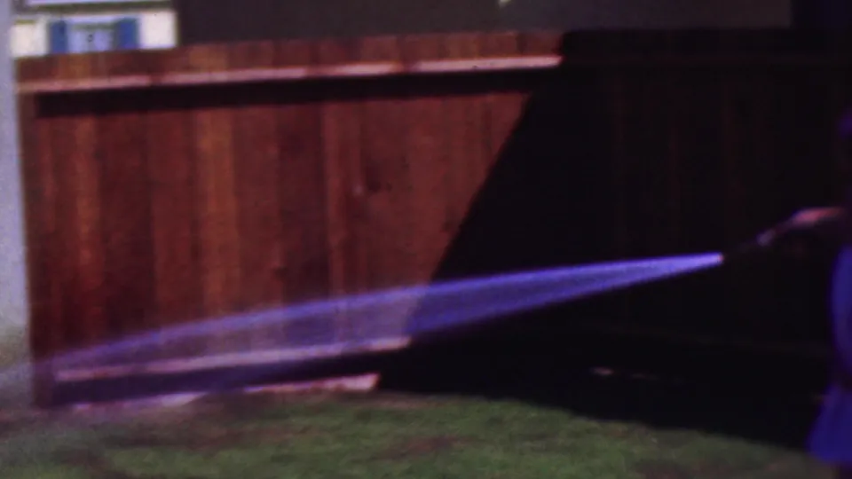 The hose stream fans across the wooden fence and low grass in the Cleveland home-movie scene, with the hose holder mostly outside the frame at the right.