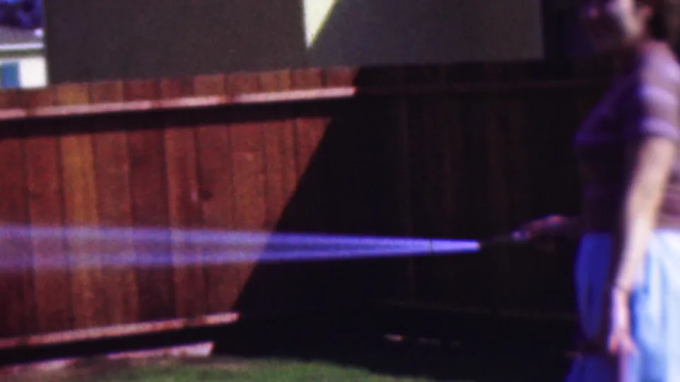 A young woman in a striped top and light skirt stands at the right edge holding a garden hose, sending a bright stream of water across the wooden fence in the 1953 Cleveland backyard footage.
