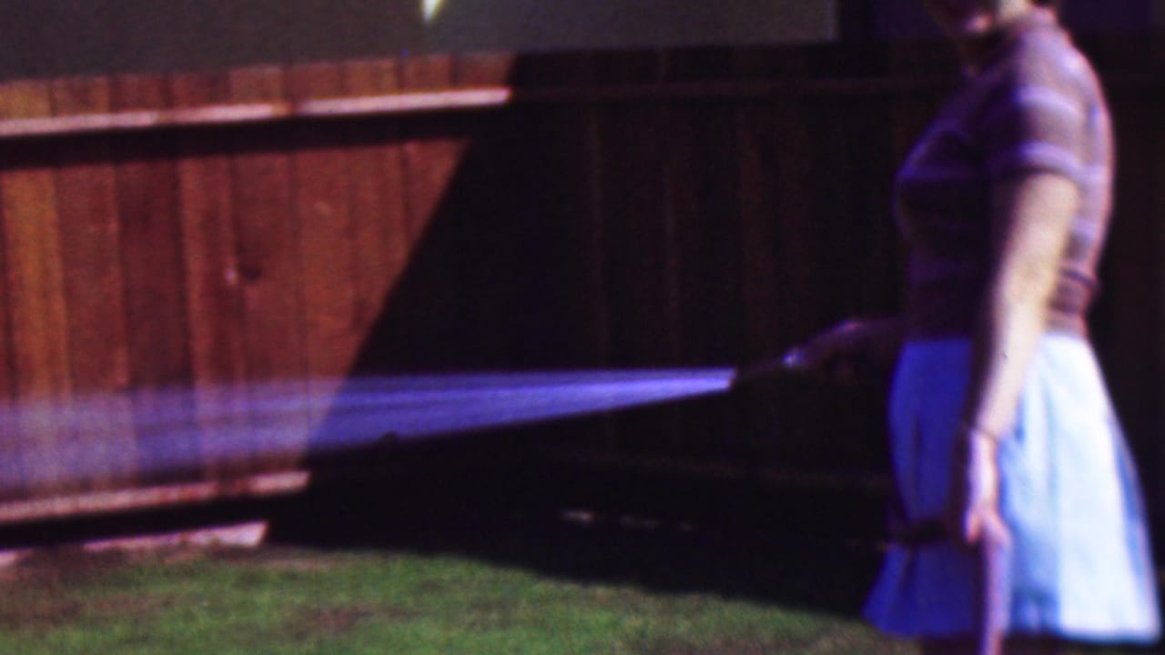 1953 Cleveland home-movie frame showing a woman in a short-sleeved shirt and light skirt watering grass beside a wooden fence.