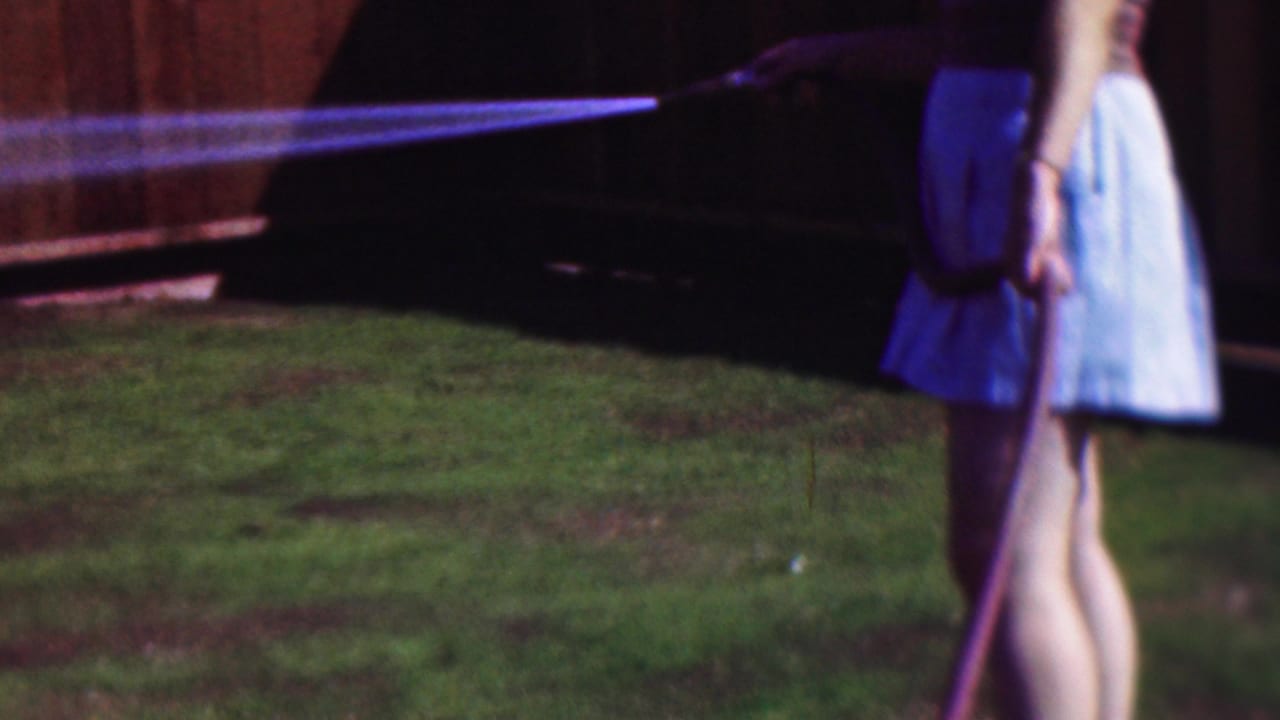 Woman holding a hose as water sprays across grass near a wooden fence.