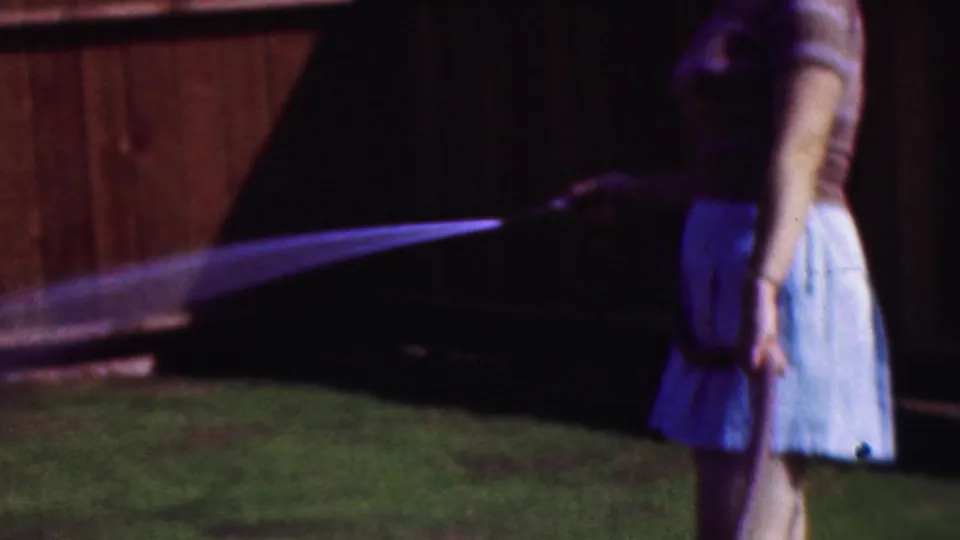 Soft 8mm frame from Cleveland in 1953 showing a woman watering a backyard lawn with a visible hose stream and fence behind her.